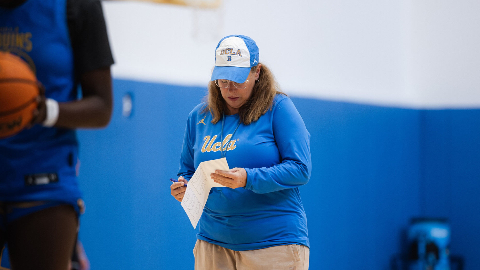 Coach Cori Close looks down and writes on her practice plan with a look of focus on her face. She stands inside the Mo Ostin Basketball Center, with blue padded walls behind her.